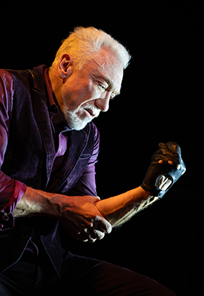 Patrick Page wearing a purple shirt and black vest, sitting and leaning forward looks at the black leather fingerless glove on his clinched left hand as his right hand grips the left arm's elbo